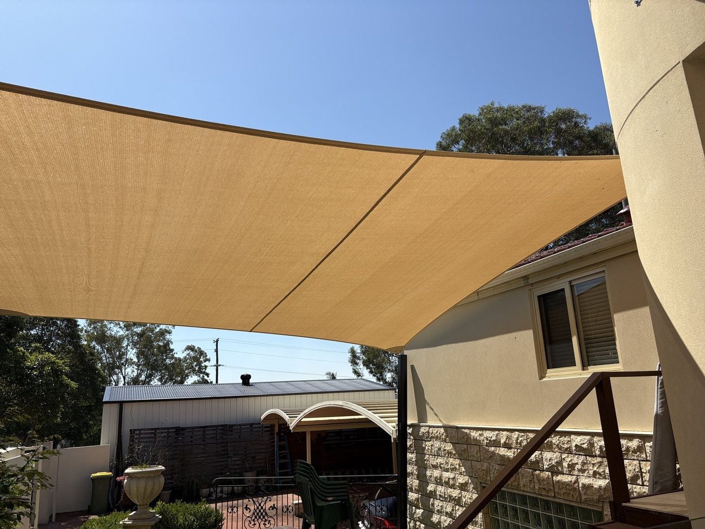 Two sand shade sails angled over a paved backyard beside a rendered home