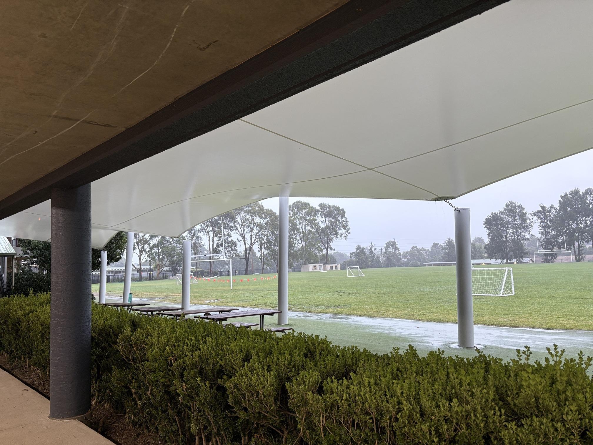 A white waterproof membrane canopy over seating beside a sports field on a rainy day